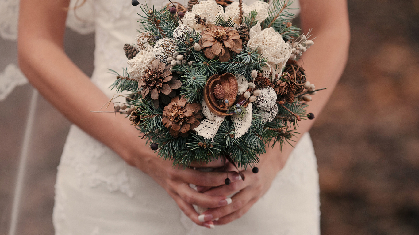 A person in a white dress holding a rustic, natural-themed bouquet with pinecones, dried flowers, lace, and greenery.