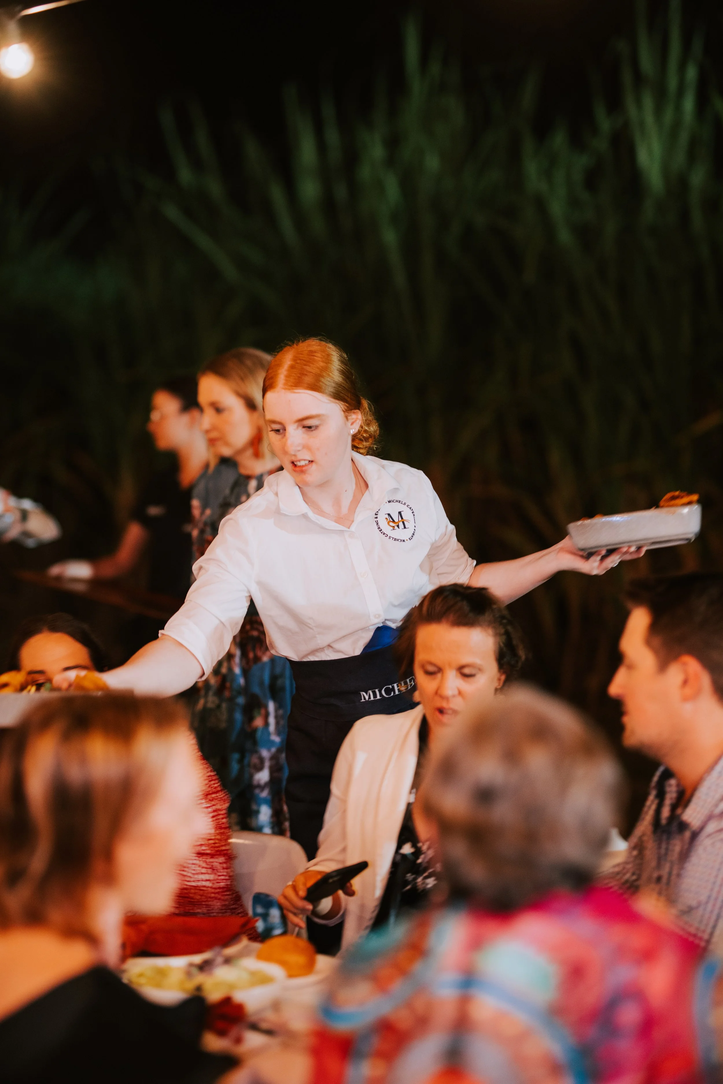 Waitress serving food at a crowded dinner event at night with a dark outdoor setting and greenery in the background.