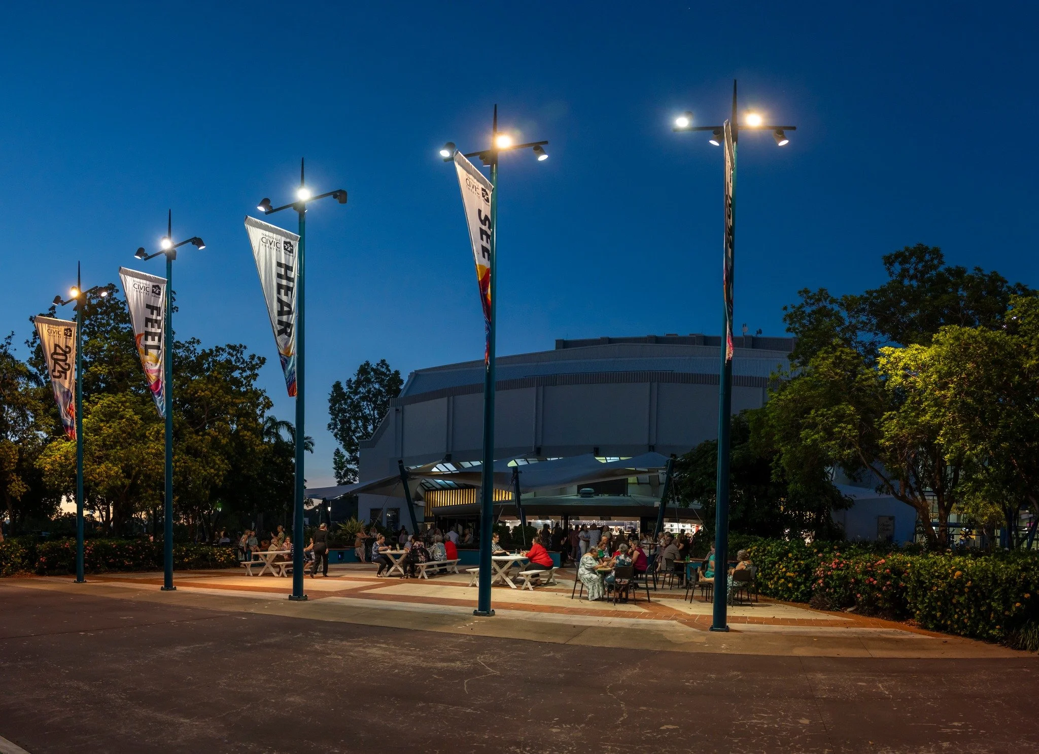 People sitting at tables outside a building during the evening at a civic event, with flags and trees in the background.