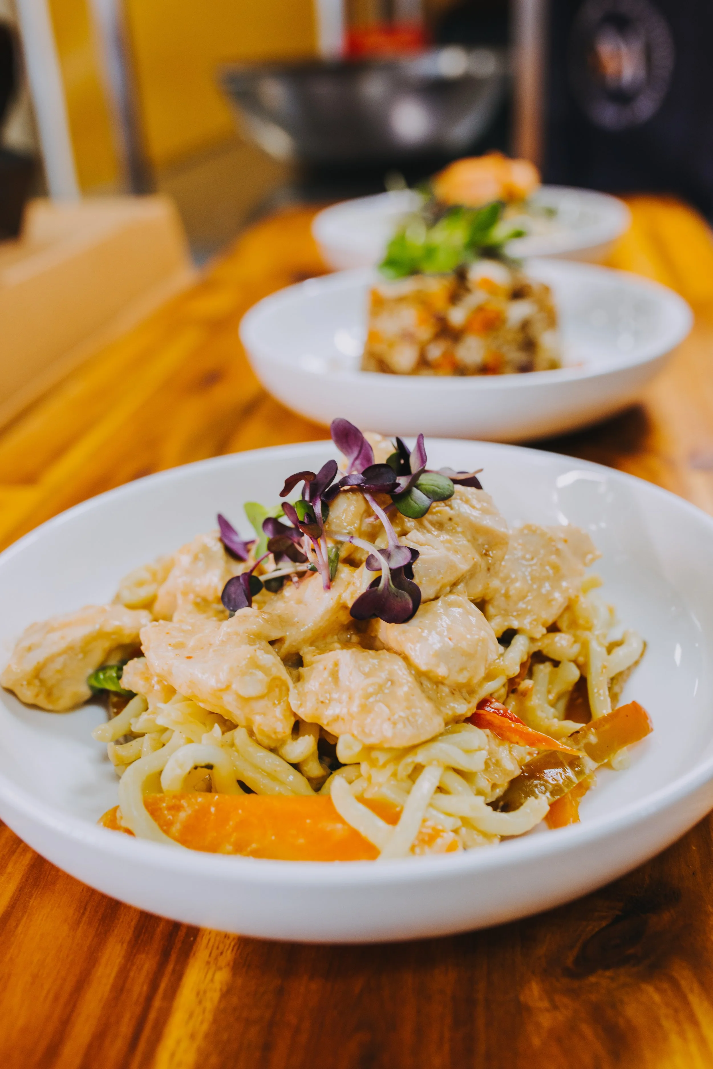 Close-up of a bowl of creamy pasta with vegetables and microgreens, with more bowls of food in the background on a wooden table.