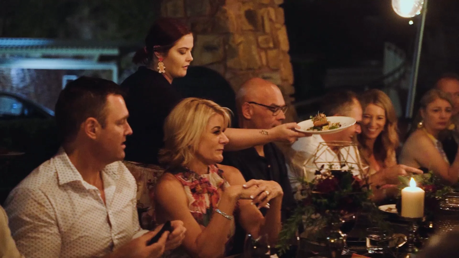 People gathered around a table at a dinner event, with one woman serving food, candlelit setting, and a rustic brick wall in the background.