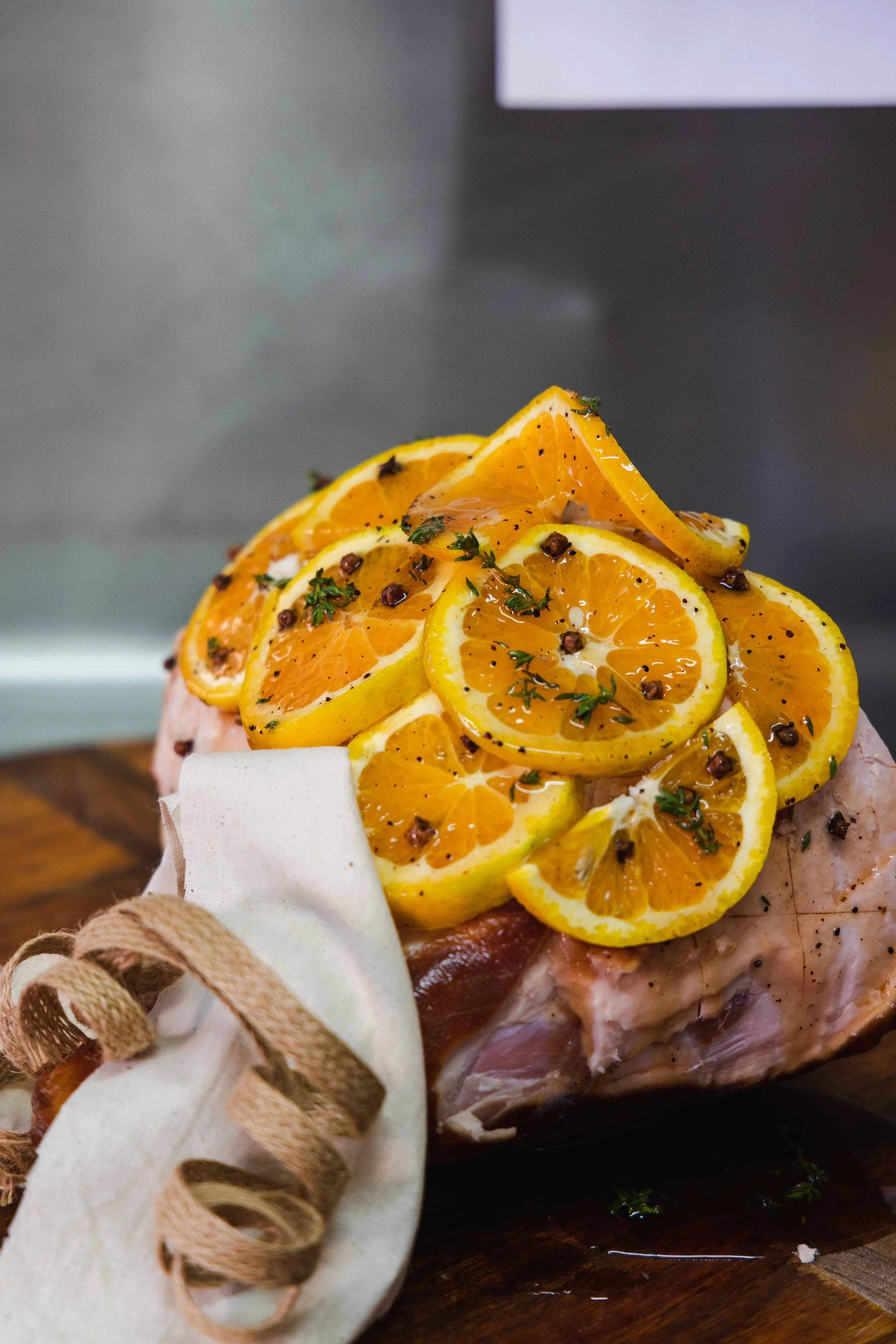 A raw or roasted ham garnished with orange slices, black pepper, and herbs, decorated with a ribbon and placed on a wooden surface.