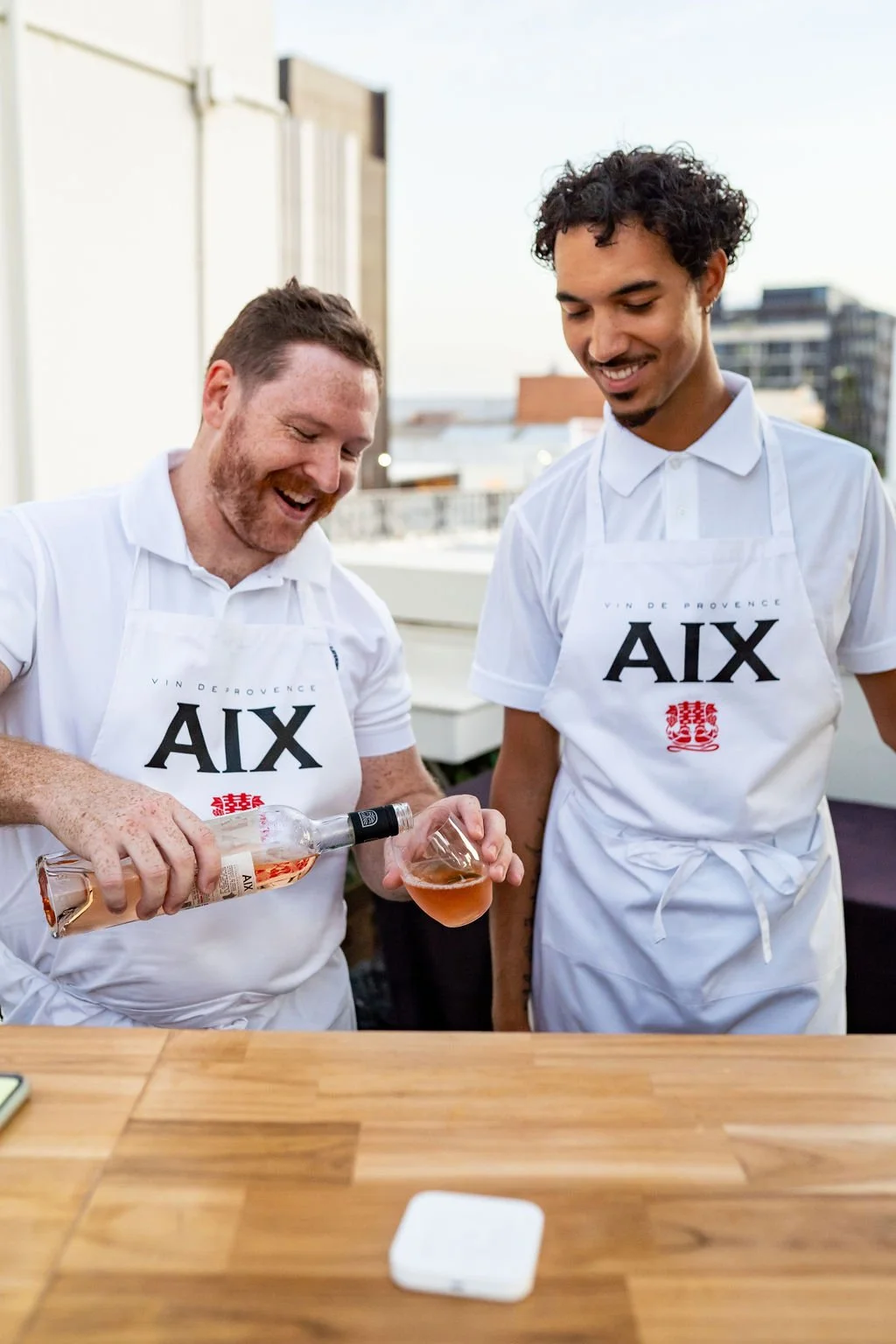 Two men in white aprons pouring and preparing drinks on a rooftop bar with city buildings in the background.
