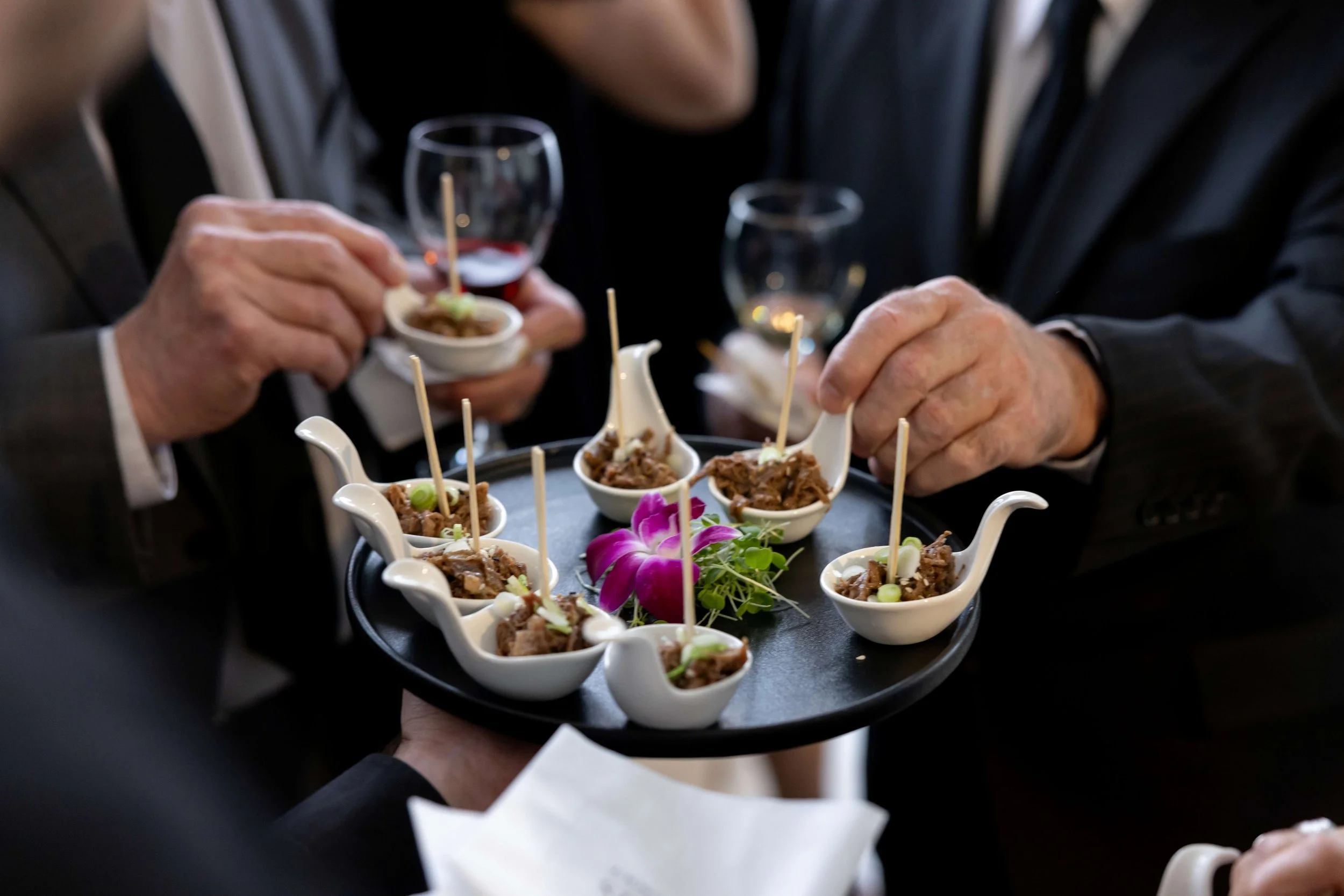 Close-up of a tray holding small bowls of meat with skewers, garnished with flowers and green onions, being served at a formal event.