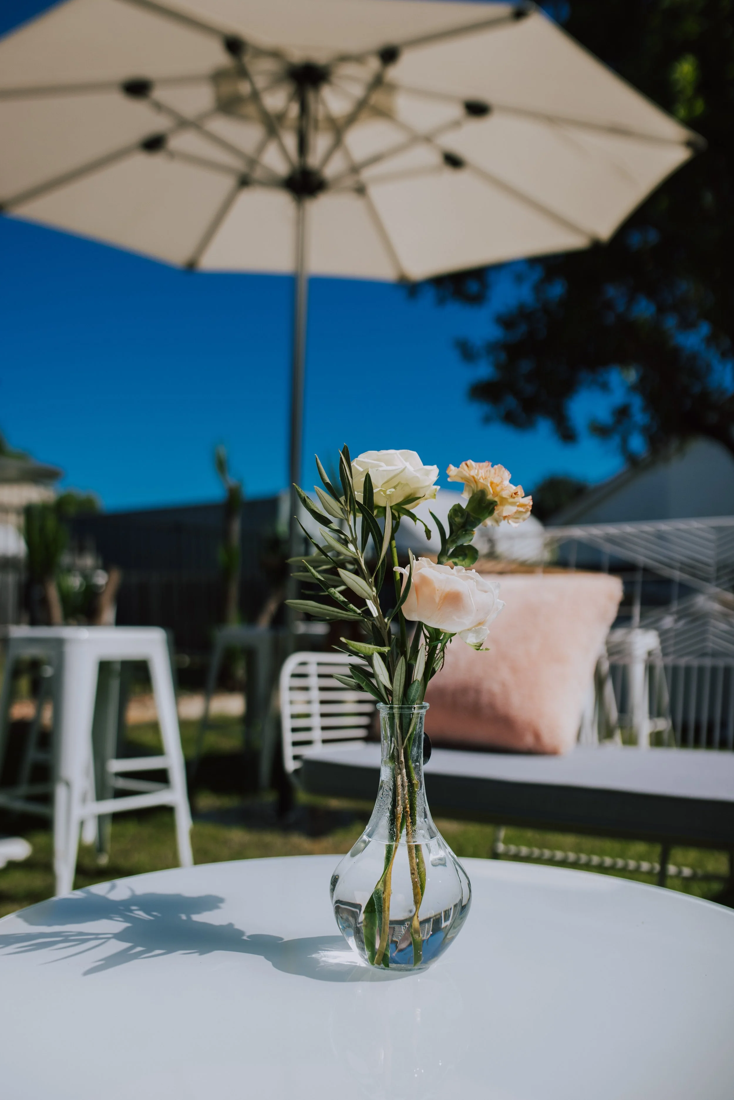 A clear glass vase with white and light pink flowers, placed on a white table outdoors under a white patio umbrella.