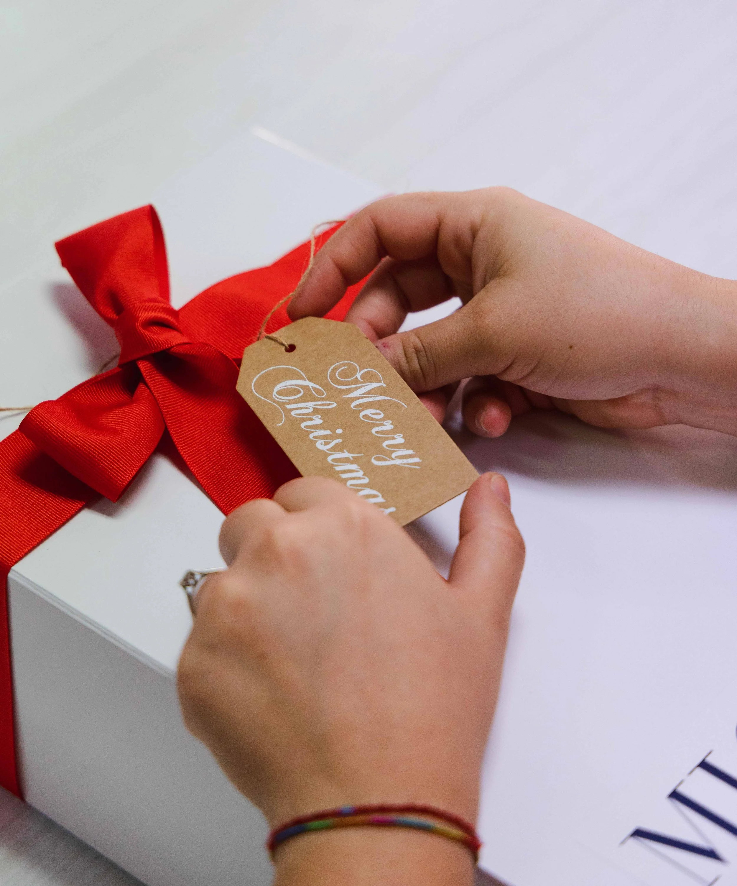 Person holding a gift tag with handwritten words, red ribbon decoration, white background, hand wearing colorful rubber bracelets.