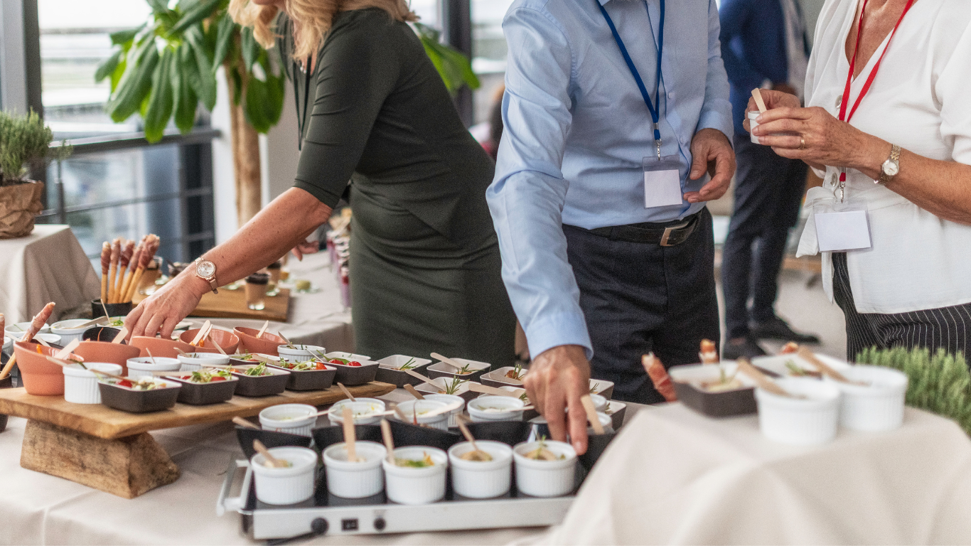 People serving themselves food at a buffet table with small bowls containing appetizers or dips, in a bright room with large windows and plants.