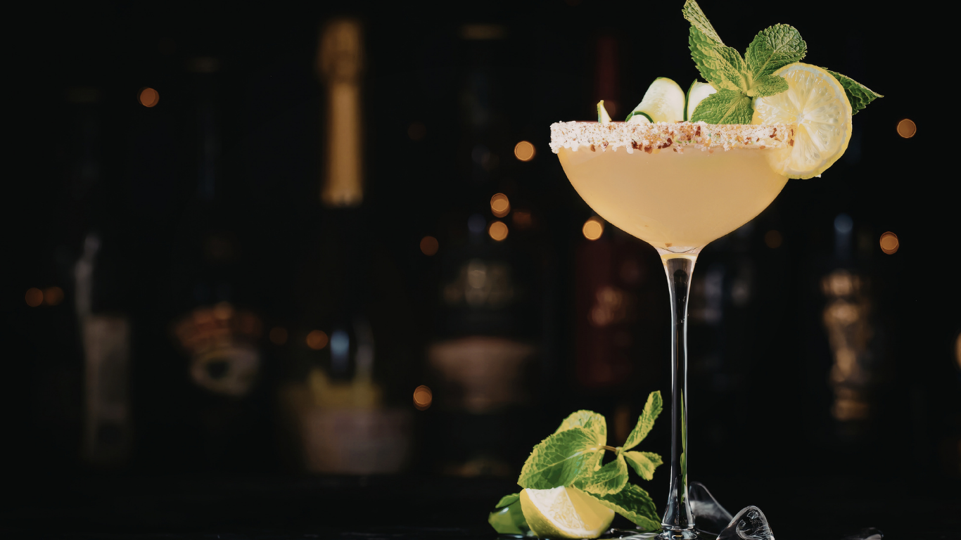 A yellow cocktail garnished with lemon slices, mint leaves, and cucumber slices in a cocktail glass on a dark background with blurry lights.