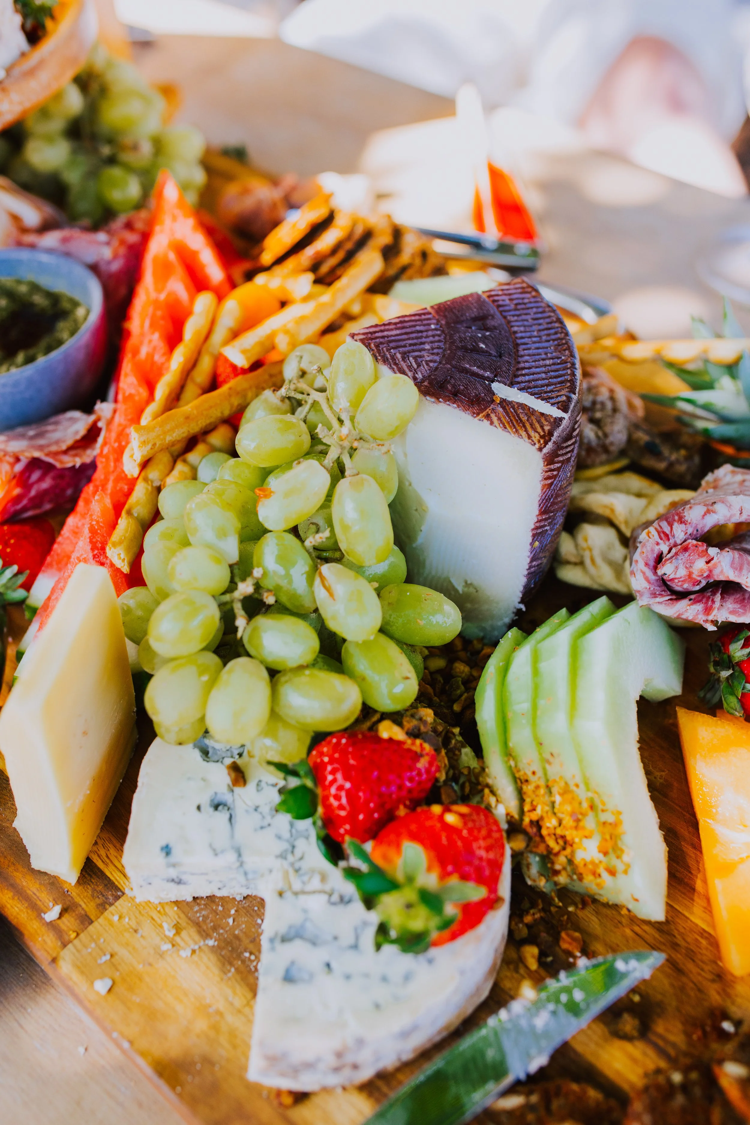 Cheese platter with grapes, strawberries, sliced vegetables, and various cheeses on a wooden board.