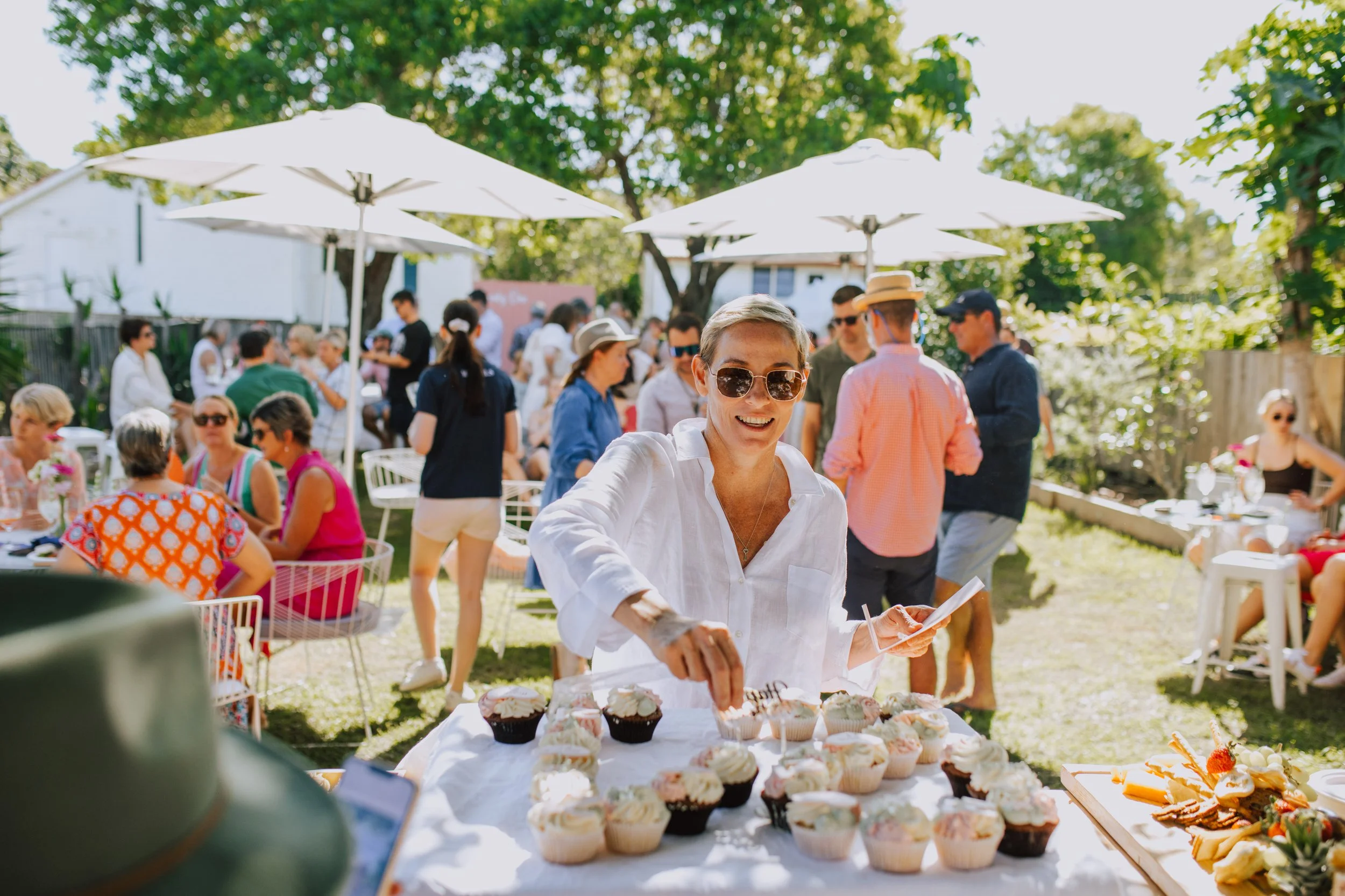 A woman with sunglasses and a white shirt serving cupcakes at an outdoor gathering under white umbrellas, with many people socializing in a lush green backyard.