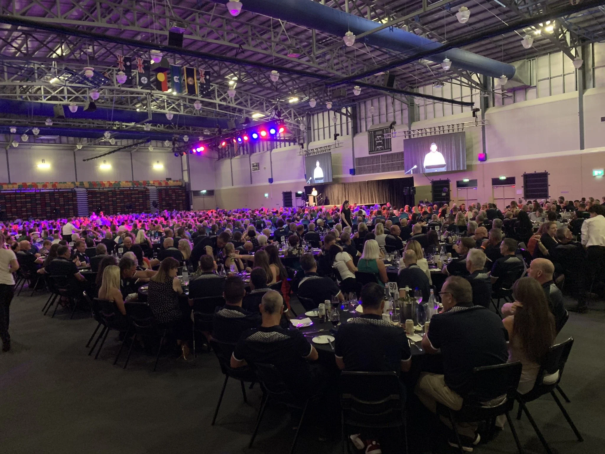 A large indoor event with many people seated at tables, watching a speaker on stage. The stage has large screens displaying the speaker and is illuminated with colorful lights. The ceiling features flags hanging from the rafters.