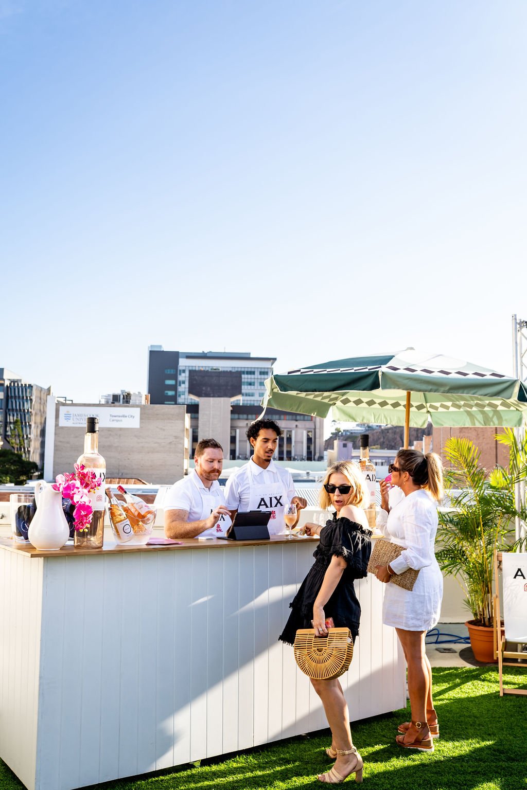 People standing at an outdoor bar with umbrellas and potted plants, city buildings in the background, on a sunny day.