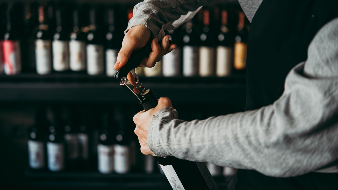A person opening a wine bottle with a corkscrew in front of a wine shelf.