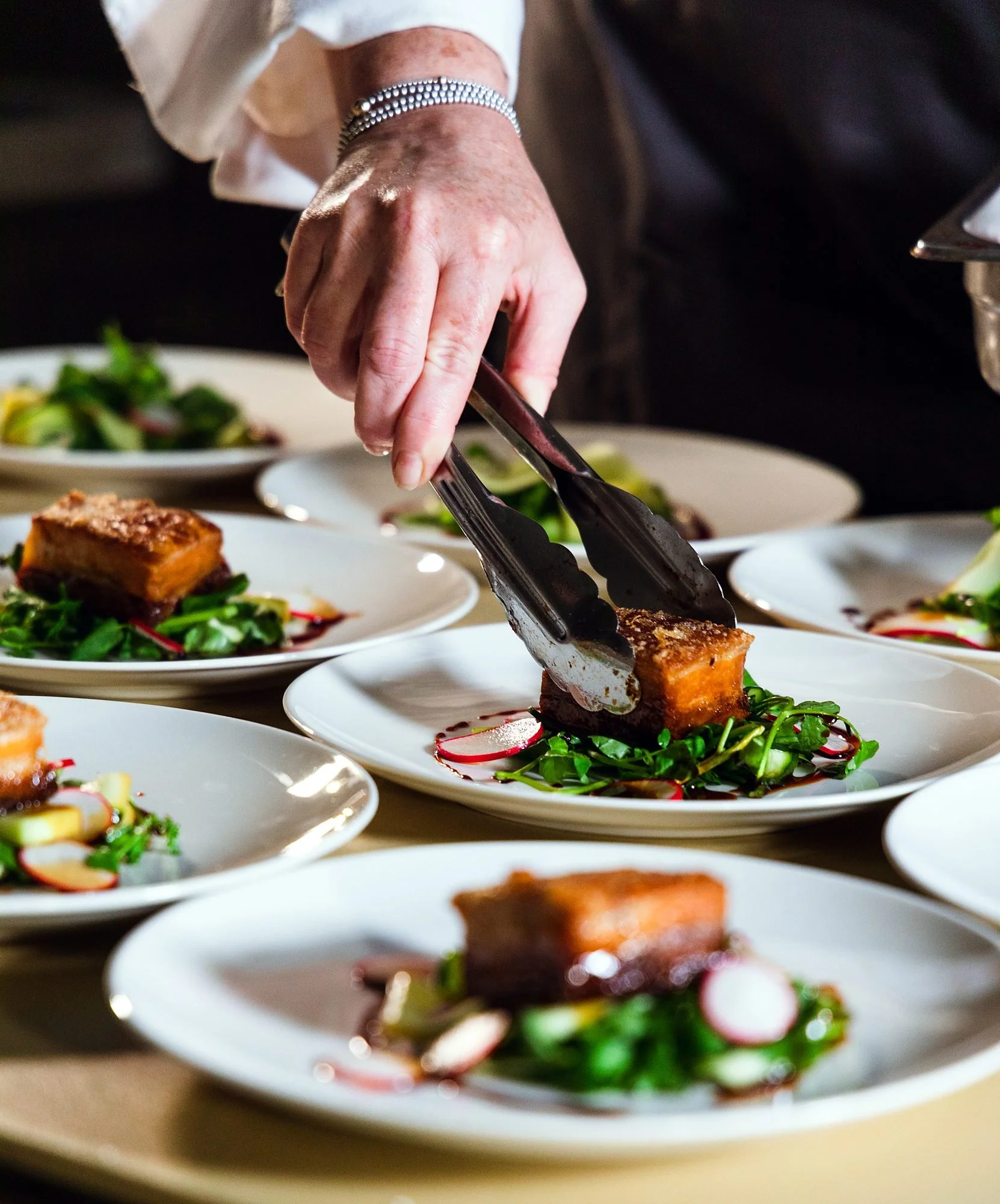 Chef plating pork belly mains with tongs for a full event menu