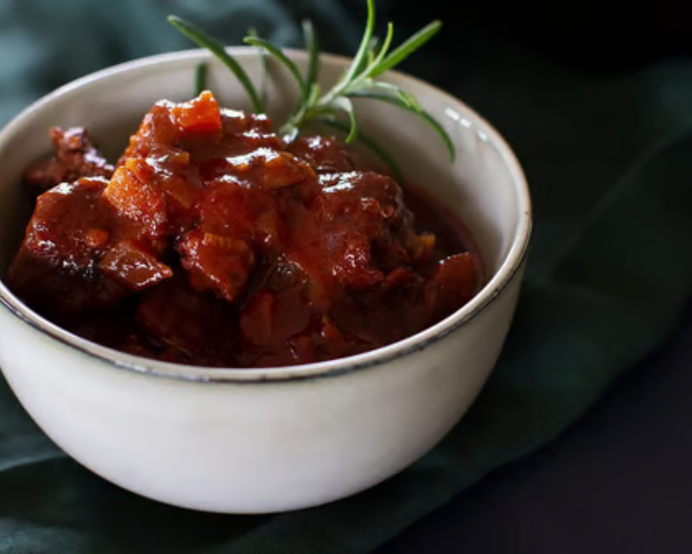 Bowl of beef stew garnished with rosemary on dark green background