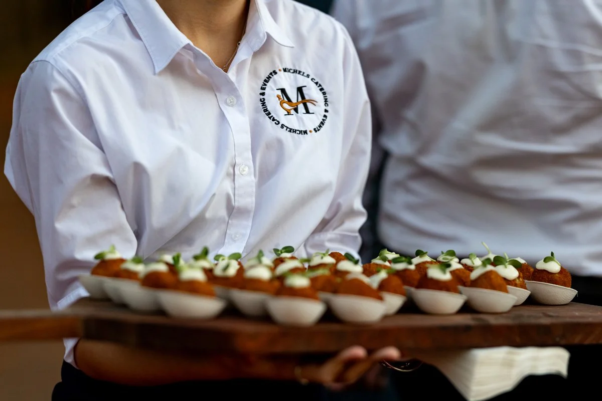 Person in a white shirt holding a wooden tray with small bowls of appetizers topped with sauce and microgreens.