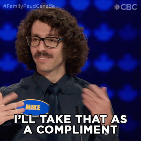 A man with curly hair, glasses, and a mustache, wearing a black shirt and tie, smiling while holding his shoulders, on a game show with the text 'Family Feud Canada' visible in the background.