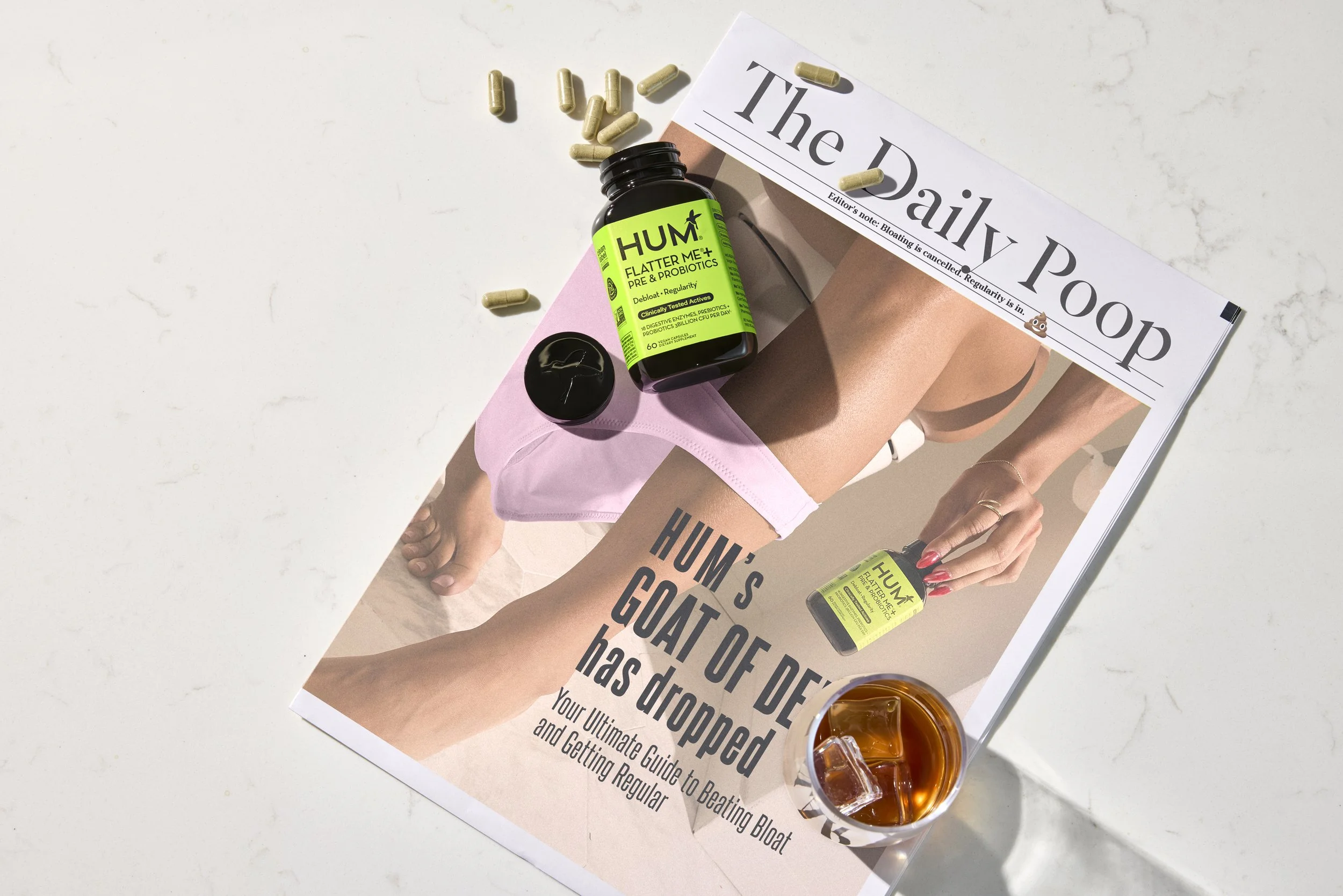 Top Prop Stylist DANA KAPUSTIN set designer and art director created a scene featuring HUM Nutrition's Flatter Me over a printed newspaper pills scattered across page. Cup of ice coffee sits next to bottle spilled open with pills scattered. 