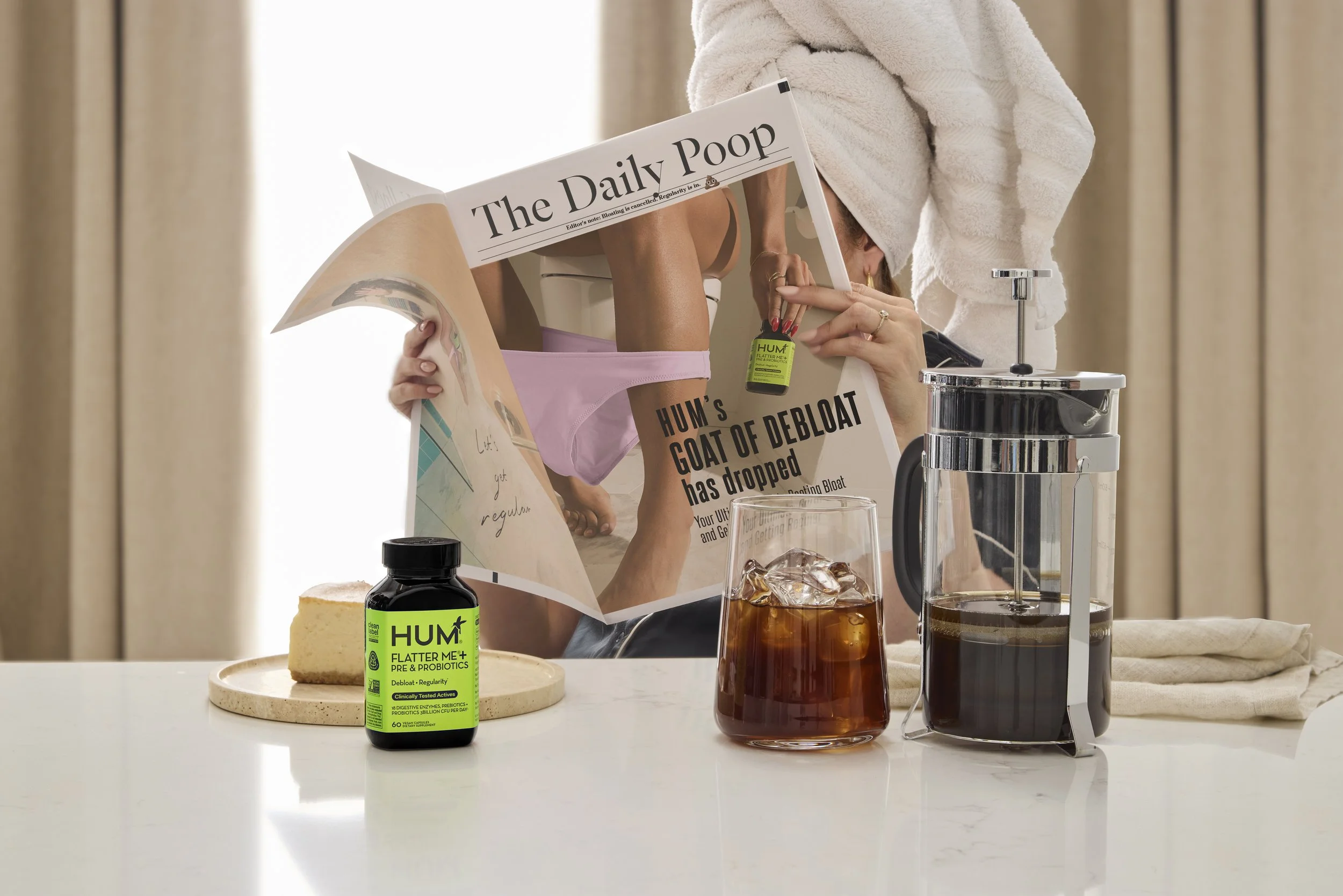 Set Designer, Prop Stylist, Art Director DANA KAPUSTIN created a fake hotel room featuring false window, drapes, and HUM Nutrition's Flatter Me bottle. Ice coffee sits next to bottle with French press on. white table top surface. 