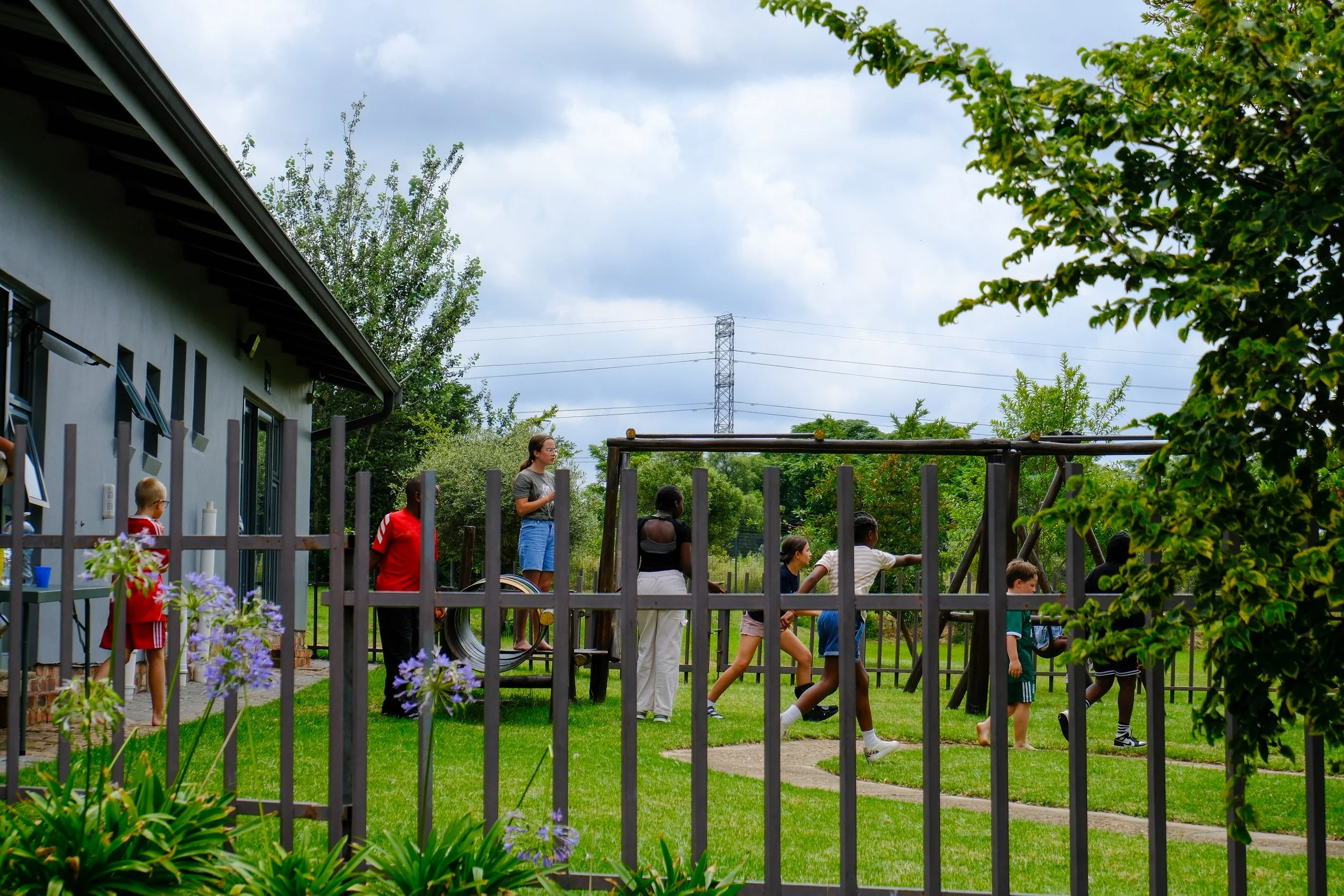 A group of children and an adult woman playing outside in a backyard with green grass and trees, seen through a black metal fence, in front of a house with gray walls and a garden with plants and flowers.