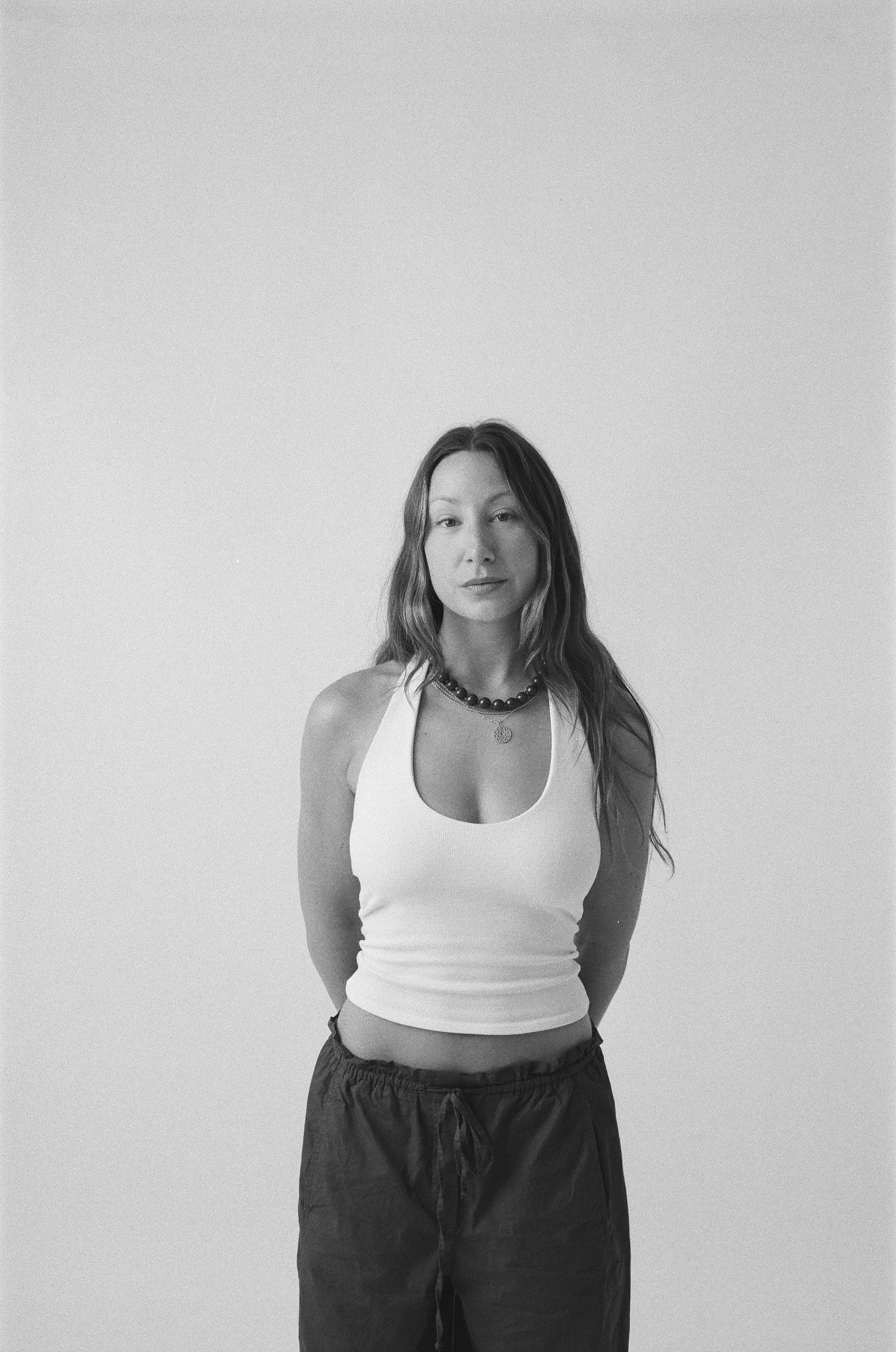Black and white photo of a woman with long wavy hair, wearing a white tank top and dark drawstring pants, standing against a plain wall.