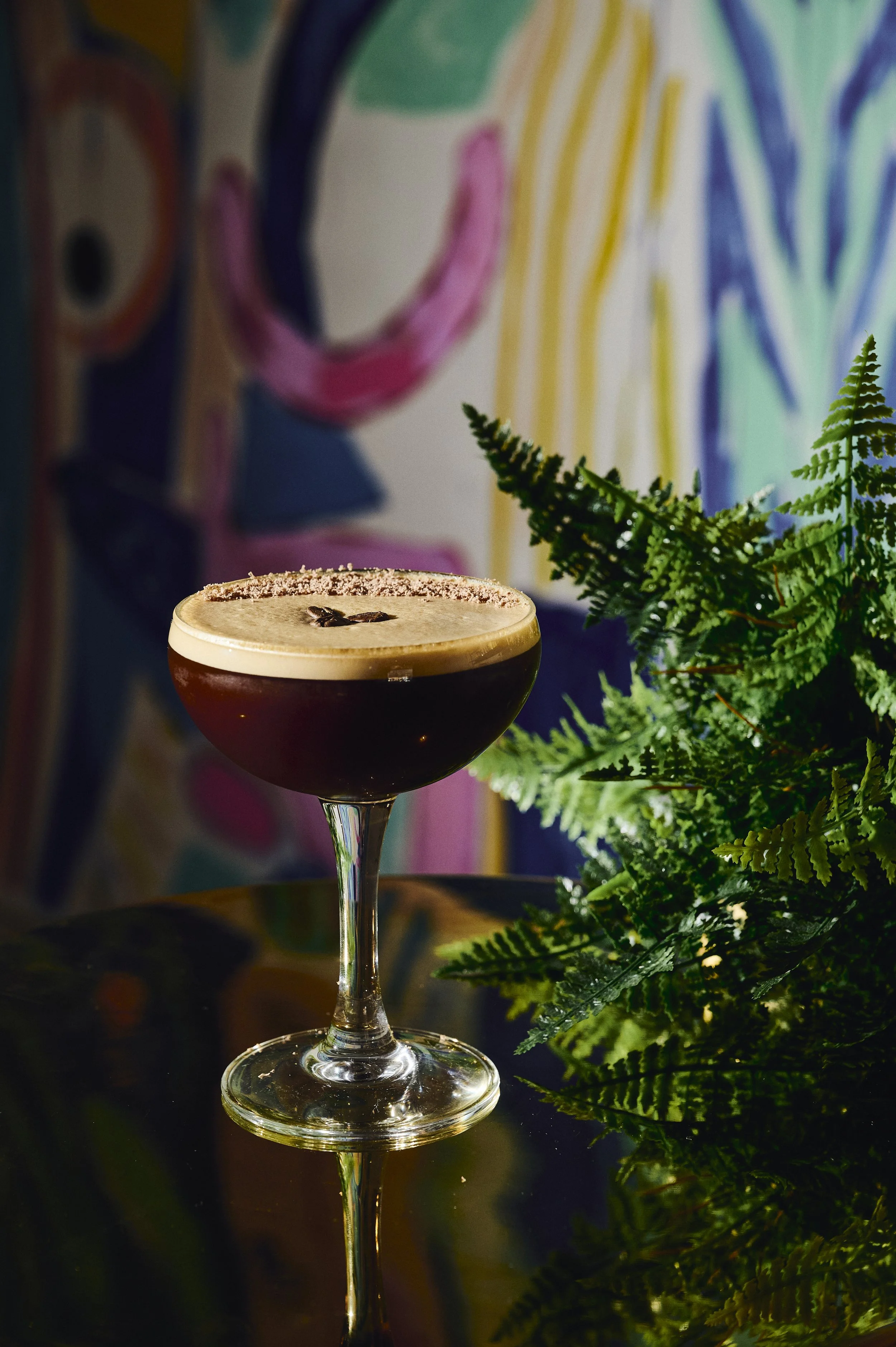 A dark coffee cocktail in a coupe glass garnished with a star anise, placed on a reflective black surface with green ferns and a colorful abstract background.