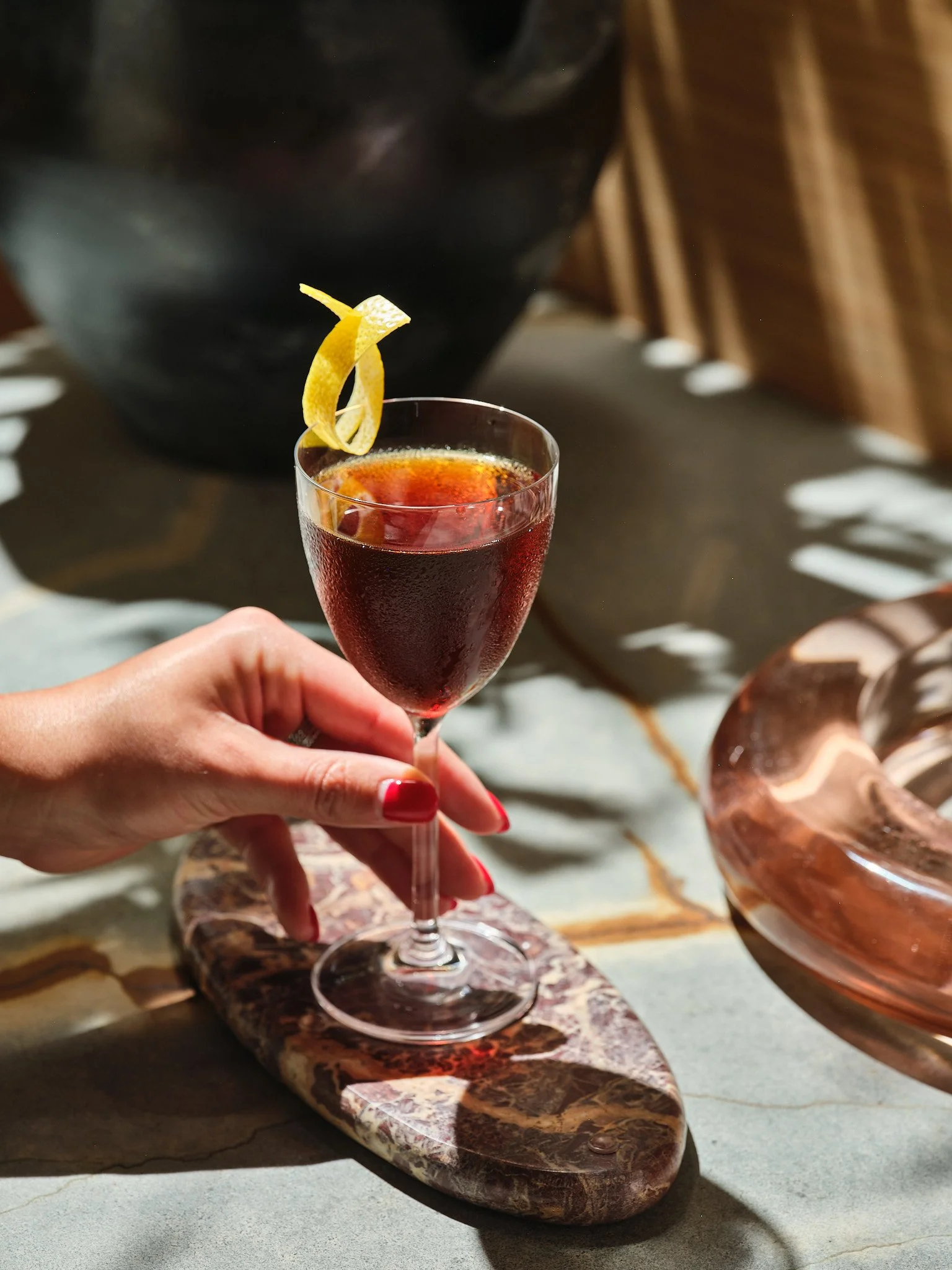 A hand with red painted nails holding a glass of red wine or cocktail with a lemon twist garnish, placed on a marble surface with decorative objects in the background.
