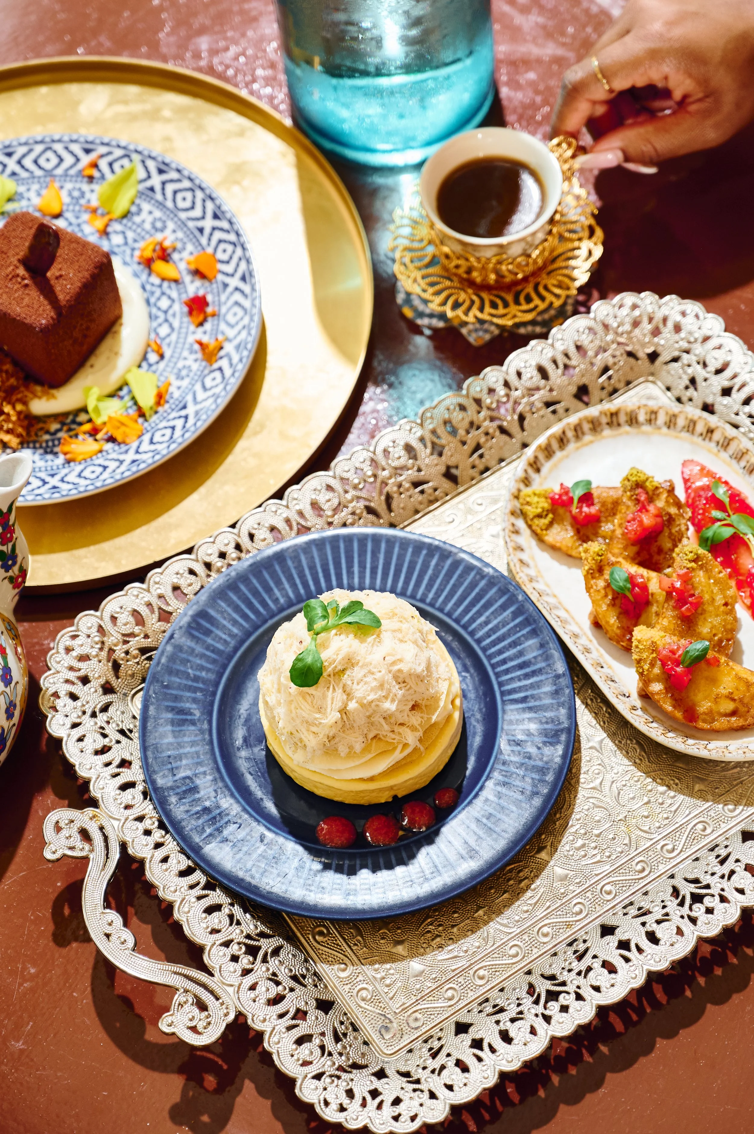 Assorted desserts and coffee on a decorative table. There is a chocolate cake, an ice cream dessert with microgreens, and fried pastries topped with pomegranate seeds and herbs. A person's hand holding a cup of coffee is visible.
