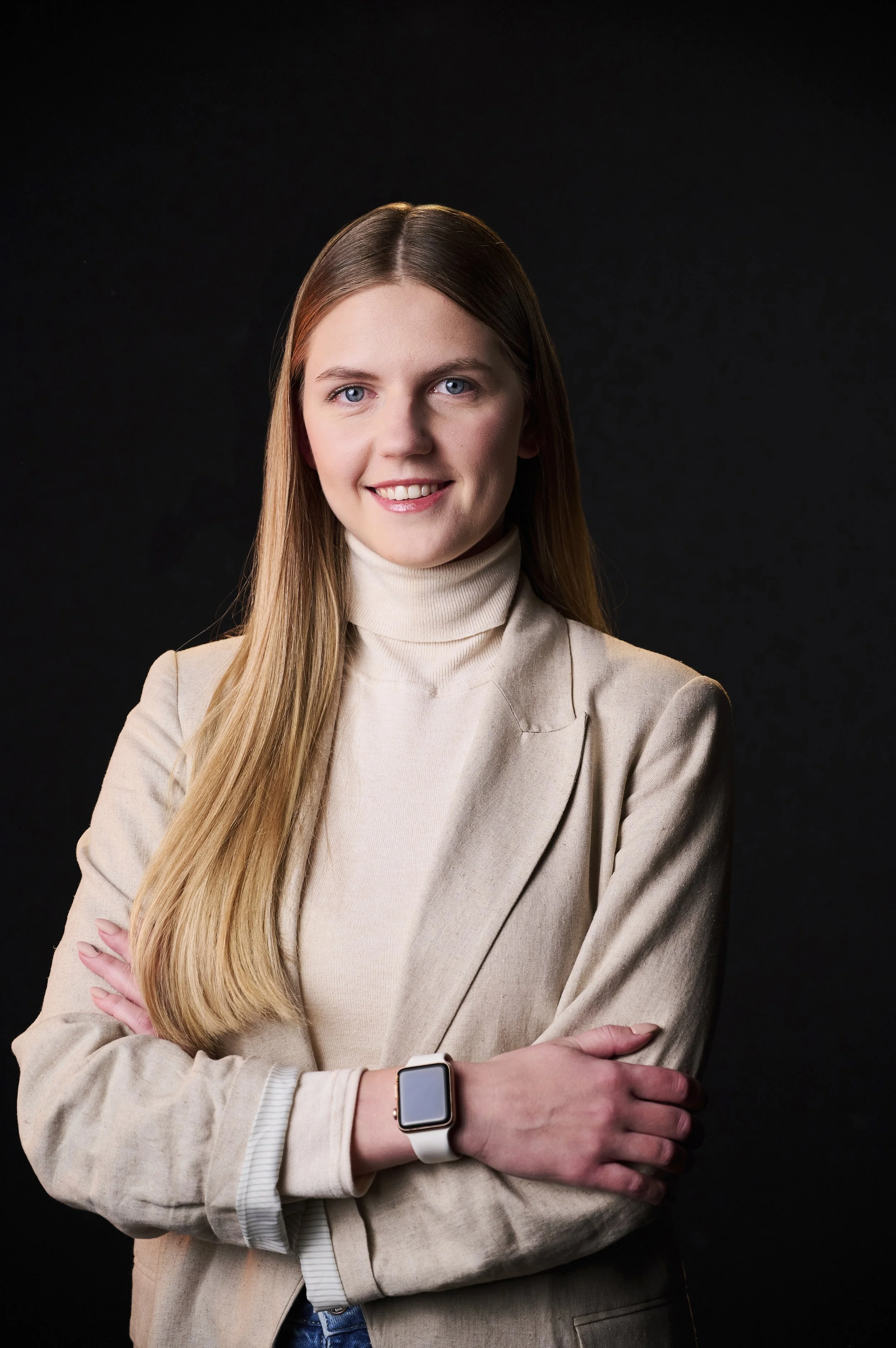 Young woman with long blonde hair wearing a beige blazer and turtleneck, smiling with arms crossed, against a dark background.