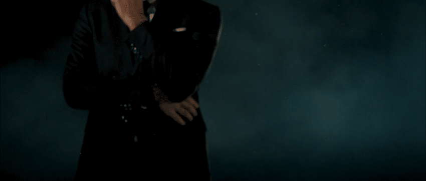 A person dressed in black clothing standing against a dark background with clouds or fog.