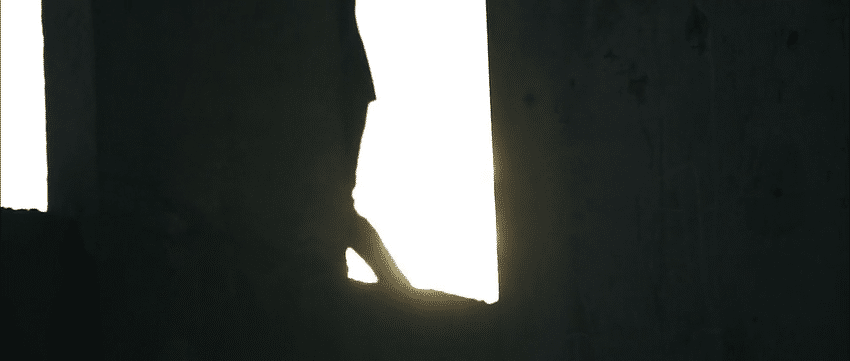 Silhouette of a person peeking through a narrow opening in a wall during daylight.