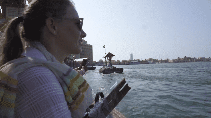 Side view of a smiling woman with sunglasses, holding a tablet, standing near a waterfront with buildings and a small boat with a flag in the background.