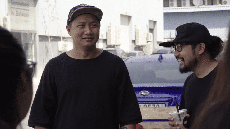 A group of people engaged in conversation outdoors, with two men in focus, one wearing a black shirt and cap, and the other with glasses, a beard, and a black cap, holding a drink.