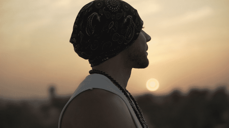 Silhouette of a man in profile wearing a bandana and necklace, standing outdoors at sunset.