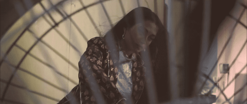 A woman with glasses, long hair, and a patterned jacket looking down, seen through a circular wire mesh or arch.