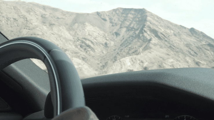 View from inside a car showing a mountainous landscape through the windshield, with a steering wheel partially visible on the left.