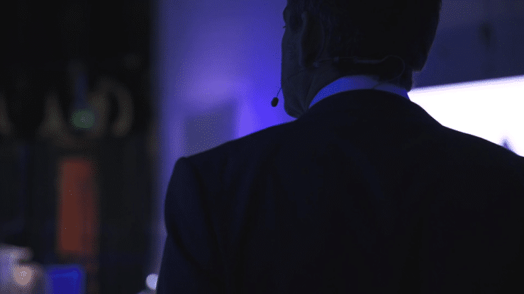 A man in a suit wearing a headset with a microphone, facing away in a dark environment with purple lighting.