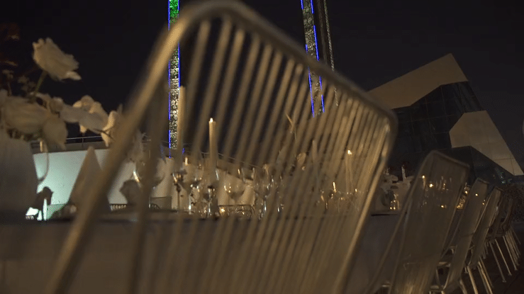 Empty outdoor dining area at night with metal chairs and table settings, illuminated by nearby lights.