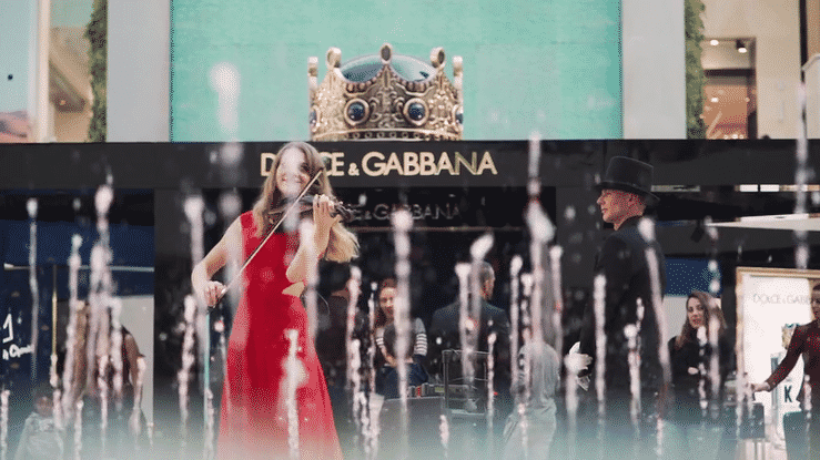 Two people, a woman in a red dress playing violin and a man in a black suit and bowler hat, standing among water fountains at a shopping mall, with a Dolce & Gabbana store in the background.