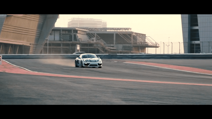 A silver sports car speeding around a racetrack with modern buildings in the background.