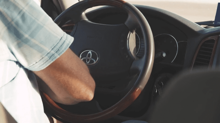 Close-up of a person with a white sleeve driving a vehicle with an orange interior, steering wheel, and dashboard.