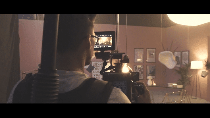 A person operating a camera on a tripod in a dimly lit room decorated with framed photos, plants, and modern lamps.