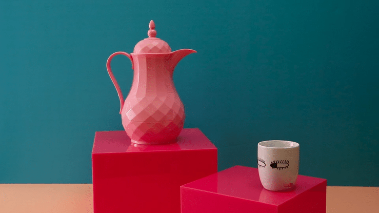A pink geometric-style teapot and a white cup with black lash illustrations on pink pedestals against a teal background.