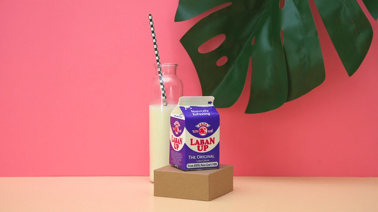 A carton of LABAN UP milk on a small block, with a glass bottle of milk and a black-and-white striped straw behind it, and a large green monstera leaf in the background against a pink wall.