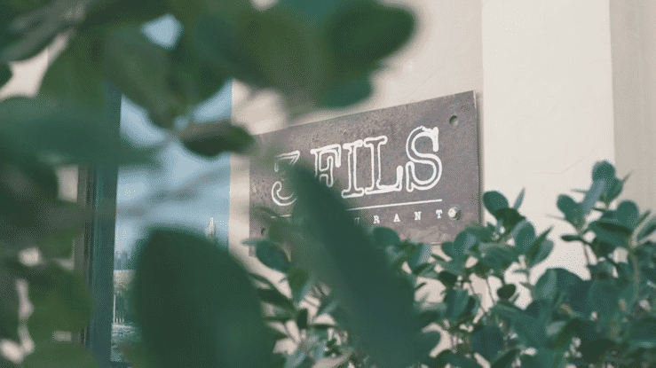 Neon sign reading 'EATS' partially obscured by green leaves in front of a light-colored wall.