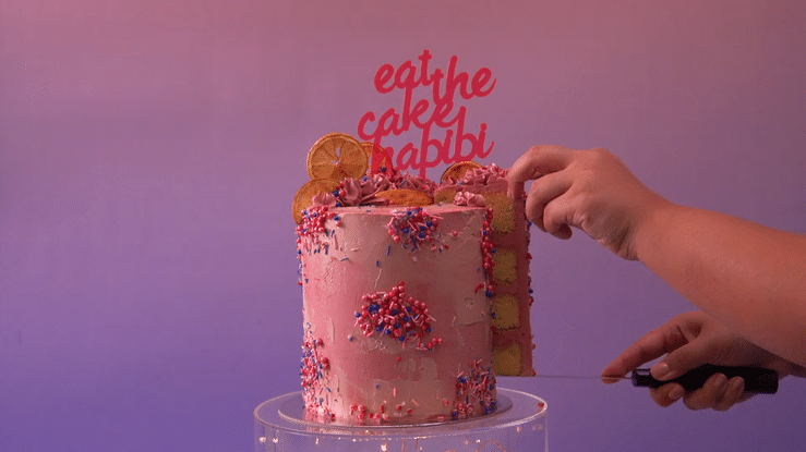 A hand is cutting a pink, decorated cake with sprinkles and slices of dried orange on top, placed on a clear stand against a gradient pink and purple background. A red cake topper reads "eat the cake, habibi".