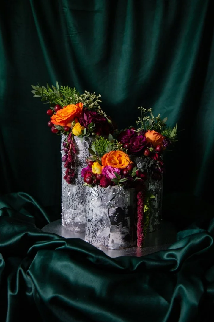 A floral arrangement with orange, yellow, and purple flowers, including roses and berries, in a cracked silver vase on a dark green satin cloth backdrop.