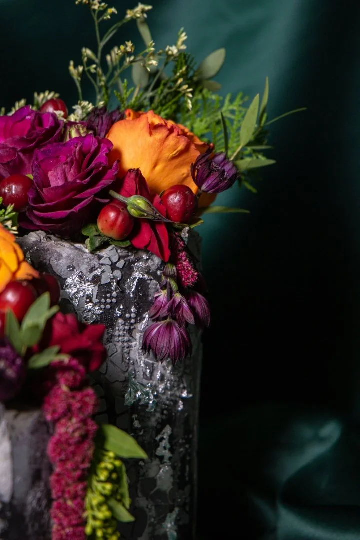 Close-up of a floral arrangement with purple roses, orange and purple flowers, red berries, and green foliage in a decorative vase against a dark background.