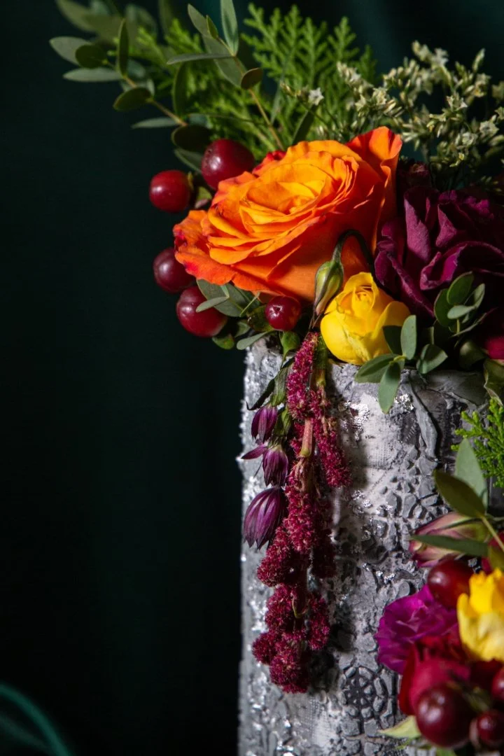 Close-up of a colorful flower arrangement in a textured silver vase, featuring orange, yellow, purple, and red flowers with green leaves and small white blooms.