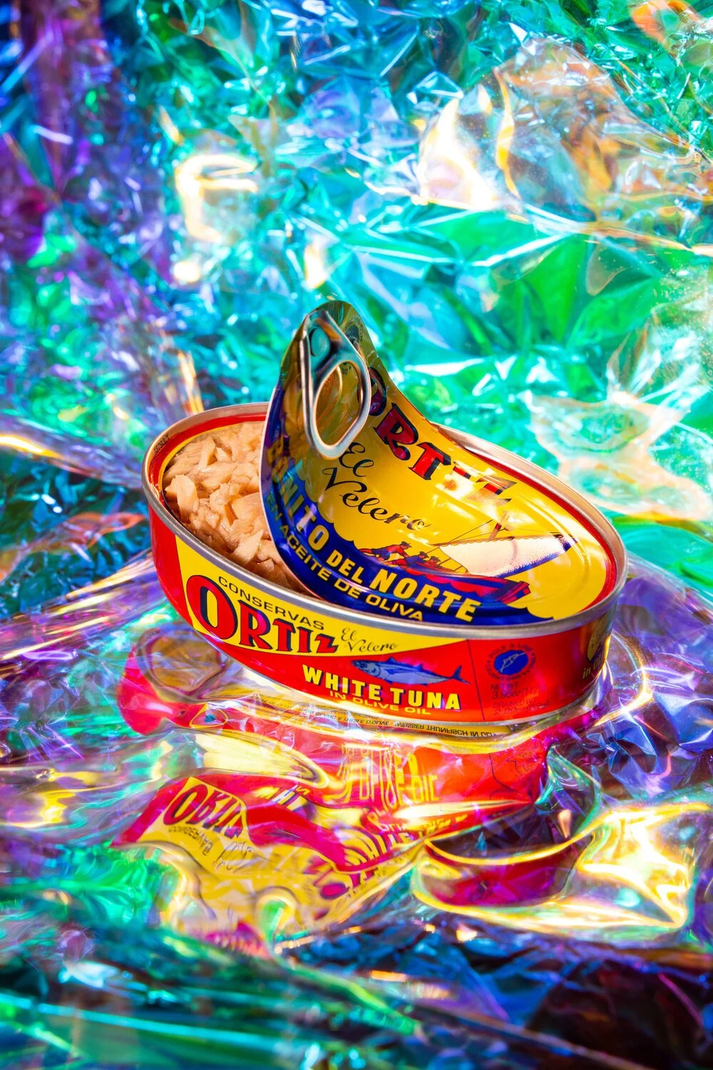 An open can of Ortiz brand white tuna in olive oil, placed on iridescent, colorful crinkled foil.